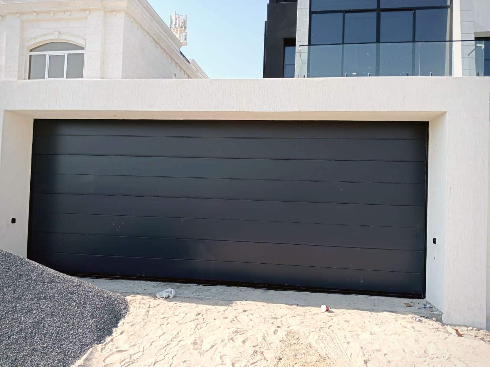 Black flush-panel insulated sectional garage door installed by BAB ALSARAY on a modern white villa in the UAE